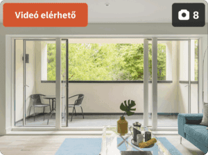 A képen egy modern nappali és erkély látható, hatalmas üvegajtóval, amely természetes fényt enged be a térbe; az erkélyen két szék és egy asztal található, zöld növényzetre néző kilátással. A bal felső sarokban narancssárga címke jelzi, hogy „Videó elérhető”, míg a jobb felső sarokban egy kamera ikon és a „8” szám utal a fotók számára.