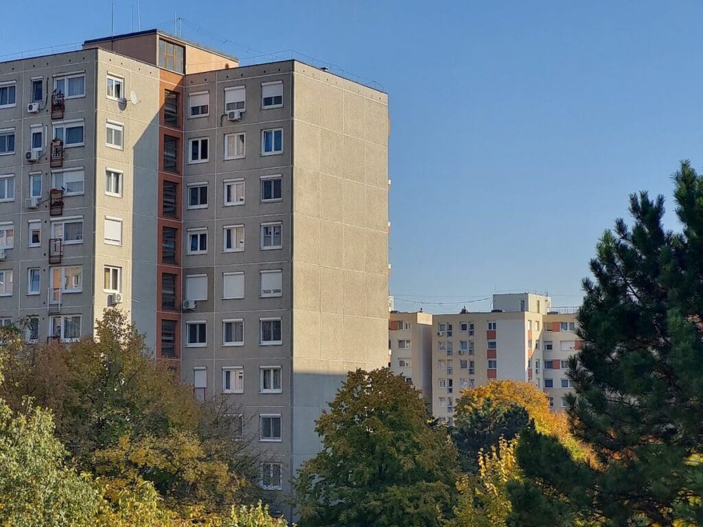 A képen többemeletes, szürke panelházak láthatók napsütéses időben, őszies lombú fákkal körülvéve. A háttérben további lakótelepi épületek tűnnek fel, a kép egy tipikus magyarországi lakótelepi környezetet ábrázol.