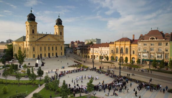 A debreceni Kossuth tér látképe a Református Nagytemplommal, környező történelmi épületekkel és szabadtéri rendezvénnyel, kora nyári napsütésben. Debrecen és Szeged környékén a legnagyobb a lakásdrágulás tempója.