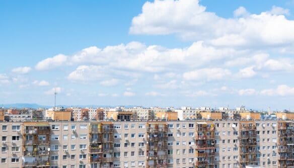 Panellakásokból álló lakótelep madártávlatból, tiszta kék ég és gomolyfelhők alatt. A képen többemeletes, erkélyes épületek sorakoznak, háttérben hegyek és városi panoráma sejlik fel.