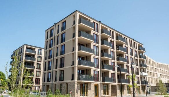 Modern, többszintes új építésű lakópark világos homlokzattal és erkélyekkel, parkosított környezetben, kék égbolt alatt. Az épület előtt rendezett köztér és fiatal növényzet látható.