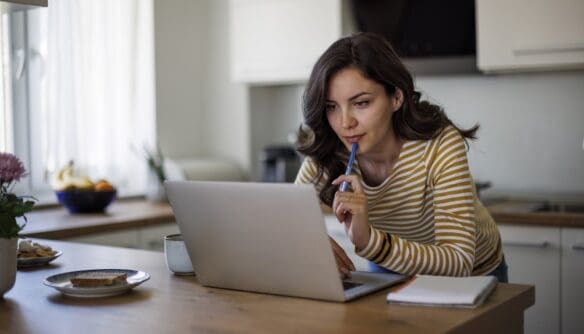Fiatal nő a konyhájában, az asztalnak támaszkodva egy laptopon keres a FIX 3%-os lakáshitel feltételeinek megfelelő otthonok között, mellette jegyzetfüzet.