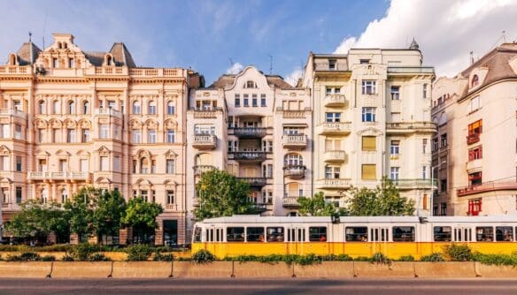 A képen egy budapesti utcakép látható, amelyen jellegzetes, díszes homlokzatú, századfordulós bérházak sorakoznak egymás mellett, előttük egy sárga-fehér villamos halad el.