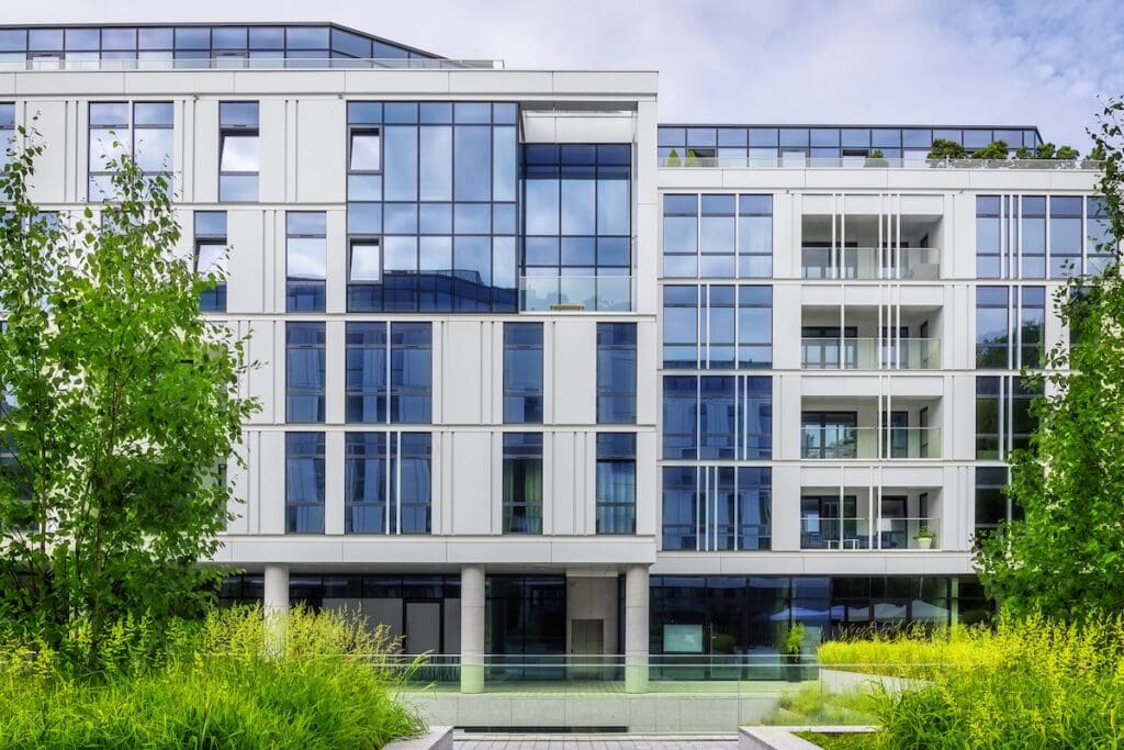 Modern új építésű társasház üveg homlokzattal és erkélyekkel, zöld kerttel körülvéve.