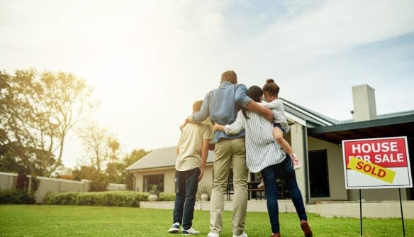 Boldog összeölelkező család hátulról, mögöttük egy családi ház. A kép jobb szélén eladó tábla jelzi, hogy az ingatlan meg volt hirdetve, a ráragasztott "Eladva" matrica alapján pedig nemrég új gazdára talált.