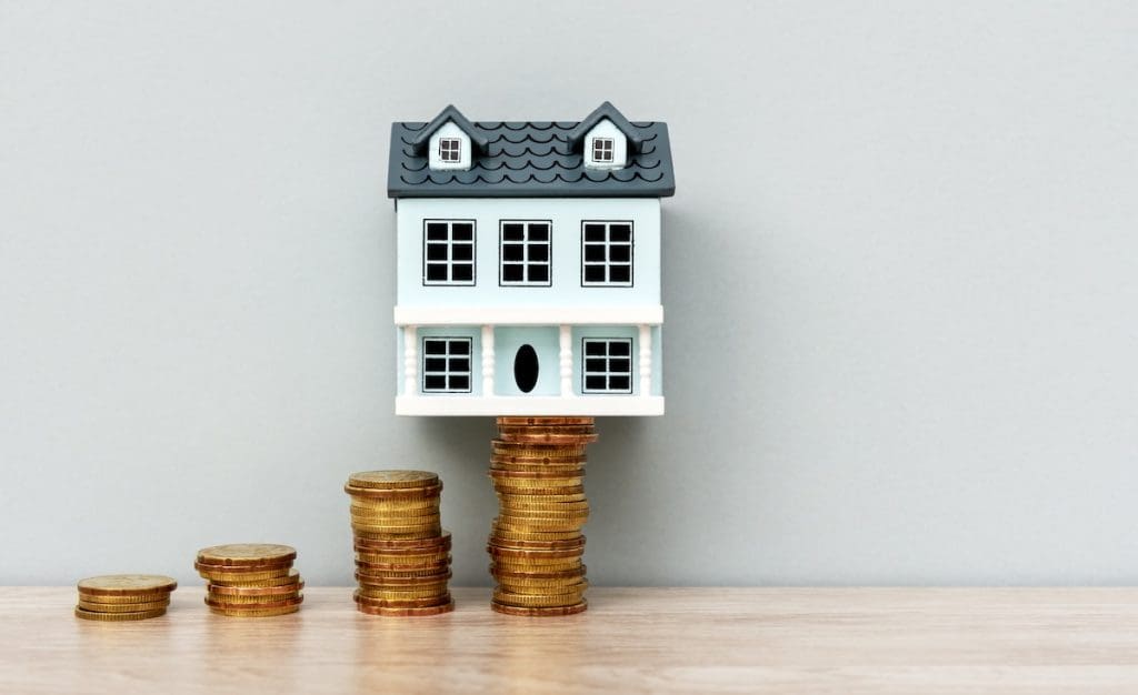 Miniatűr ház makett aranyérme oszlopokon állva, amelyek fokozatosan növekednek balról jobbra, szimbolizálva az ingatlanárak emelkedését.