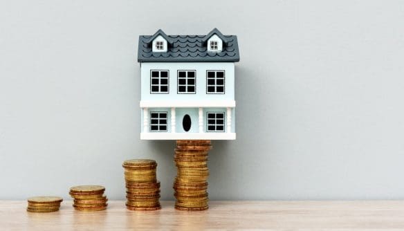Miniatűr ház makett aranyérme oszlopokon állva, amelyek fokozatosan növekednek balról jobbra, szimbolizálva az ingatlanárak emelkedését.