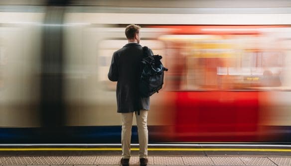 Férfi hátizsákkal vár a peronon, miközben elsuhan mellette egy piros-fehér metró szerelvény.