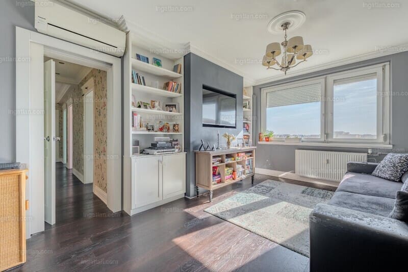 Világos, ízlésesen berendezett nappali nagy ablakokkal és városi kilátással. A falon modern TV, mellette beépített könyvespolc és szekrény található. A helyiséget elegáns csillár, szürke kanapé, dísztárgyak és növények teszik otthonossá, a falakat szürke és fehér árnyalatok uralják.