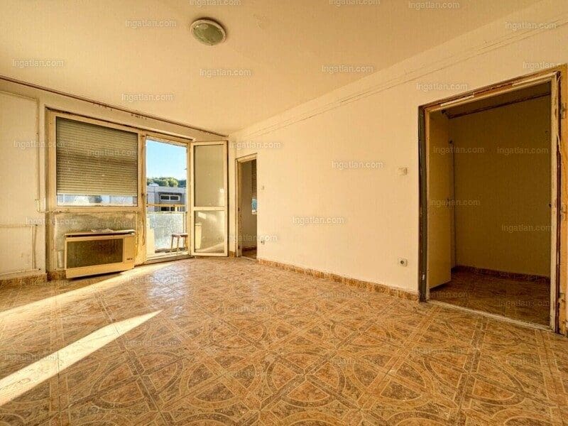 Üres, felújítandó lakás világos szobája, amelyben csempepadló, fehér falak és nagy ablakok láthatók. Az egyik falon régi gázkonvektor található, az ablakon redőny és egy ajtó vezet az erkélyre. A szoba mellett egy kisebb helyiség bejárata is látható, a tér napfényben úszik.