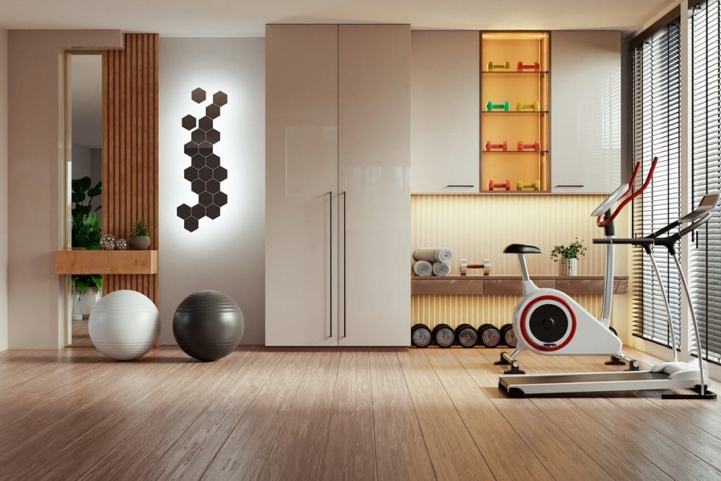 Modern, világos lakóparki fitneszterem faborítású padlóval és nagy ablakokkal. A helyiségben edzőkerékpár, futópad, kézi súlyzók, jógalabdák láthatók. A falakat beépített szekrények és hangulatos, meleg fényű világítás díszíti.