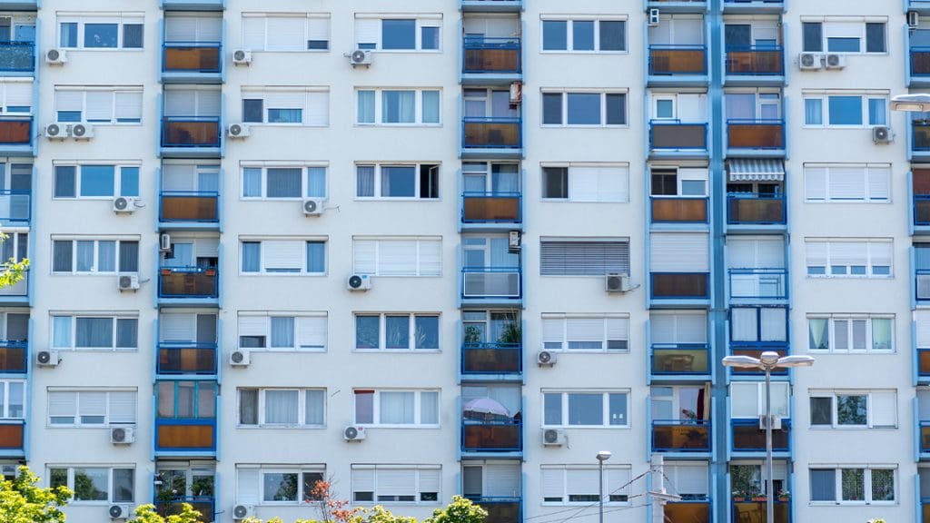 Panelház homlokzata kék erkélykorlátokkal és barna üvegfelületekkel. Az épület több emeletes, a lakások erkélyein különböző növények és napernyők láthatók. A falakon számos légkondicionáló berendezés található, az előtérben pedig néhány fa és utcai lámpa.