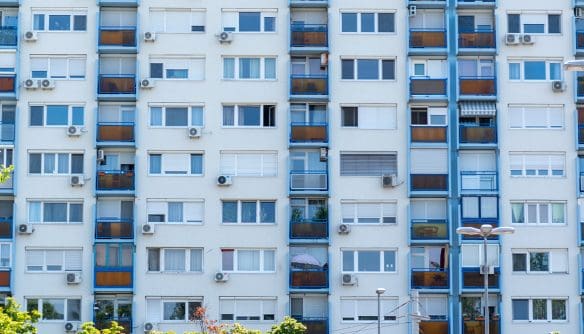 Panelház homlokzata kék erkélykorlátokkal és barna üvegfelületekkel. Az épület több emeletes, a lakások erkélyein különböző növények és napernyők láthatók. A falakon számos légkondicionáló berendezés található, az előtérben pedig néhány fa és utcai lámpa.