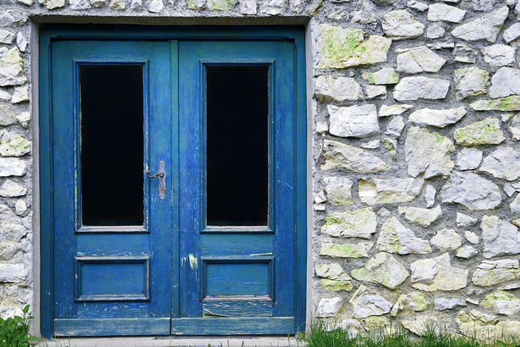 Két szárnyú, régi kék faajtó egy kőfalban. A festék több helyen megkopott, az ajtó üvegbetétes, mögötte sötét belső tér látszik. A kőfal világosszürke, helyenként zöld mohával borított, alján fű nő.