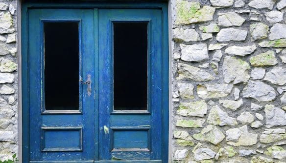 Két szárnyú, régi kék faajtó egy kőfalban. A festék több helyen megkopott, az ajtó üvegbetétes, mögötte sötét belső tér látszik. A kőfal világosszürke, helyenként zöld mohával borított, alján fű nő.