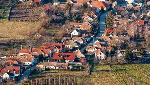 Légi felvétel egy magyar vidéki faluról, ahol családi házak és gazdasági épületek piros tetőkkel, fák és kertek között helyezkednek el. Az előtérben szántóföldek és szőlősorok láthatók, a háttérben dombos táj és további házak. A kép nyugodt, falusias környezetet mutat.