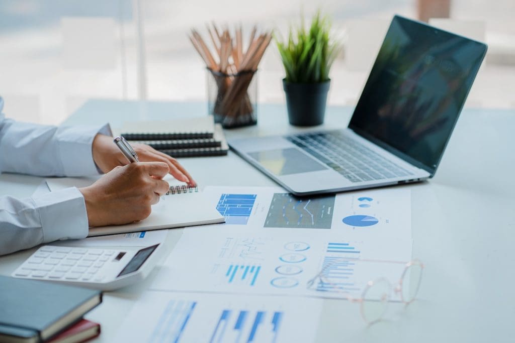 Üzletember jegyzetel egy irodai asztalnál, ahol laptop, számológép és üzleti grafikonok láthatók. A kép irodai munkát és pénzügyi elemzést ábrázol modern környezetben.”