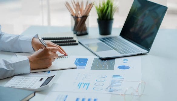 Üzletember jegyzetel egy irodai asztalnál, ahol laptop, számológép és üzleti grafikonok láthatók. A kép irodai munkát és pénzügyi elemzést ábrázol modern környezetben.”