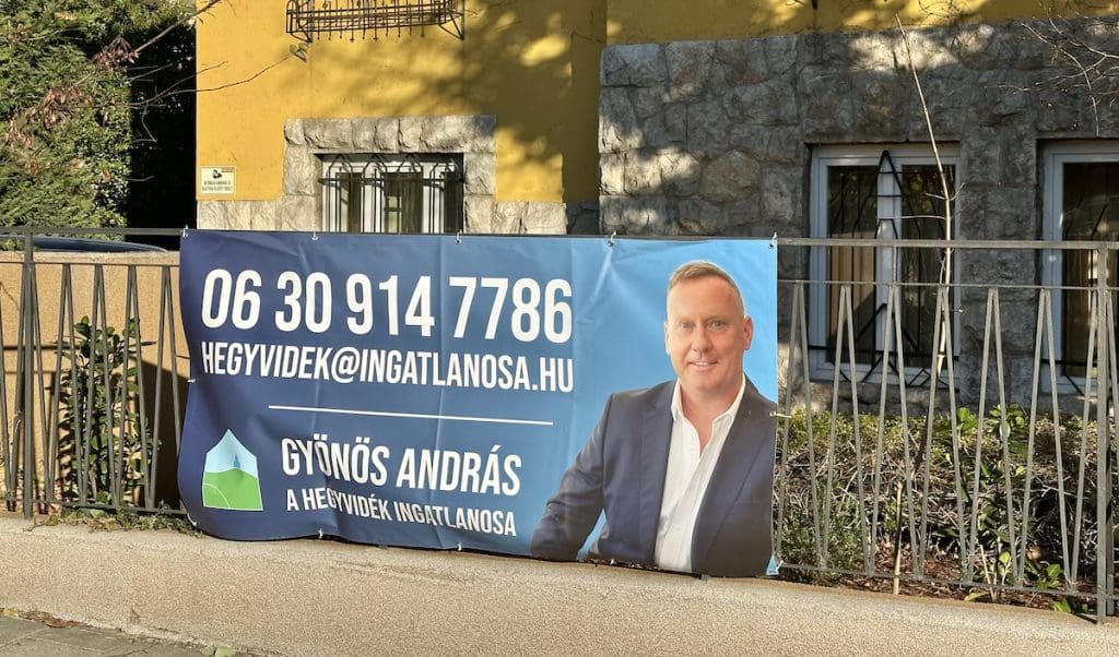 Gyönös András, az Hegyvidék ingatlanosának molinója egy ház kerítésén. A molinón András portérja mellett az elérhetősége látható. A háttérben egy sárga épület.