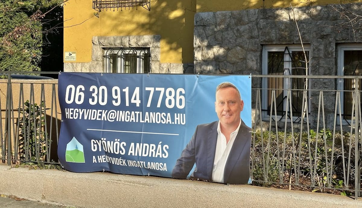 Gyönös András, az Hegyvidék ingatlanosának molinója egy ház kerítésén. A molinón András portérja mellett az elérhetősége látható. A háttérben egy sárga épület.