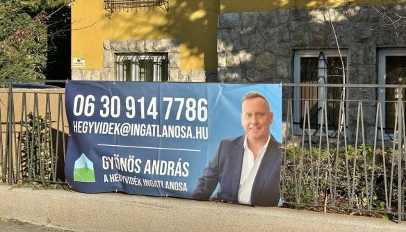 Gyönös András, az Hegyvidék ingatlanosának molinója egy ház kerítésén. A molinón András portérja mellett az elérhetősége látható. A háttérben egy sárga épület.