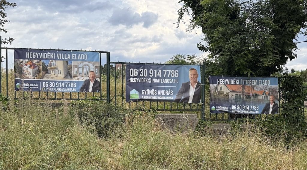 Gyönyös András plakátjai a Hegyvidék területén, egy kerítésen. A képen három egymás mellé elhelyezett plakát látható. A baloldali "Hegyvidéki villa eladó" felirattal egy házat hirdet, a középső Gyönös András portréját és elérhetőségét tartalmazza, a jobboldalin pedig egy eladó étterem fotója látható. A plakátok előtt zöld növényzet, mögötte fa és felhős ég látható.