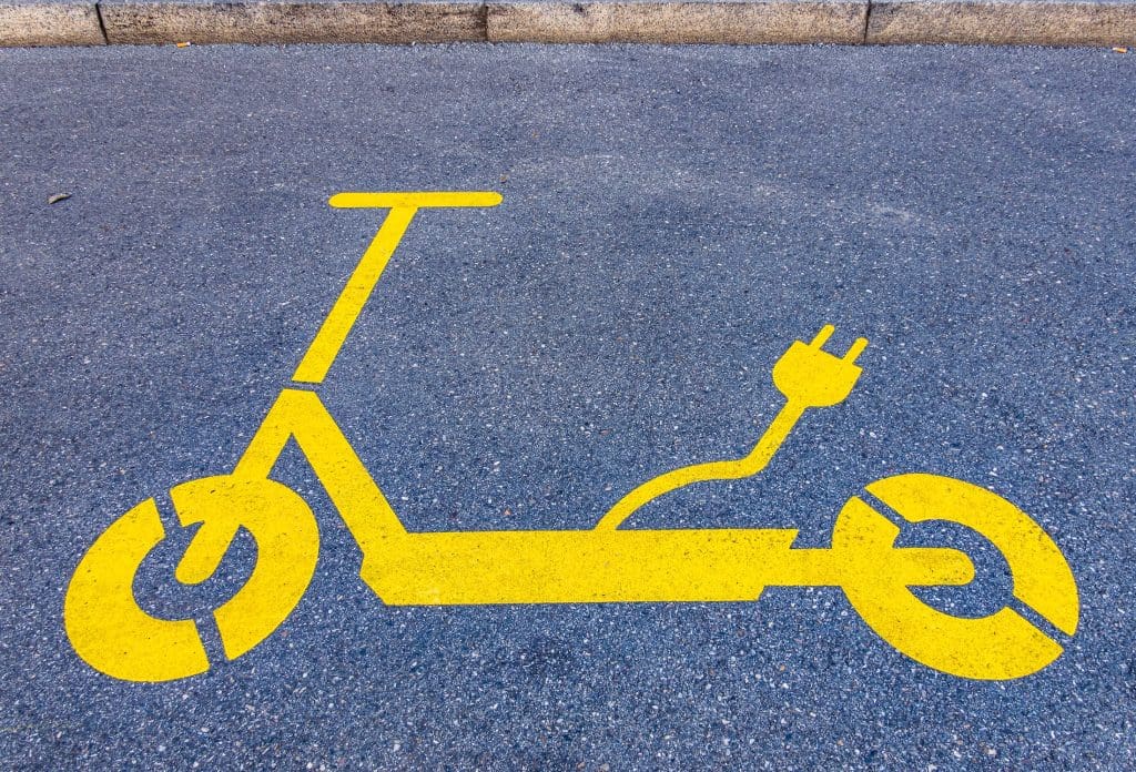 Aszfaltburkolaton látható élénksárga piktogram, amely egy elektromos rollert ábrázol. A roller hátuljából egy konnektorcsatlakozóval végződő kábel motívuma indul ki, jelezve, hogy elektromos töltőhely vagy parkolóhely megjelöléséről van szó.