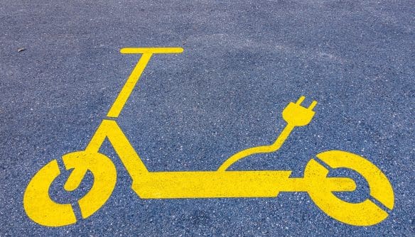 Aszfaltburkolaton látható élénksárga piktogram, amely egy elektromos rollert ábrázol. A roller hátuljából egy konnektorcsatlakozóval végződő kábel motívuma indul ki, jelezve, hogy elektromos töltőhely vagy parkolóhely megjelöléséről van szó.