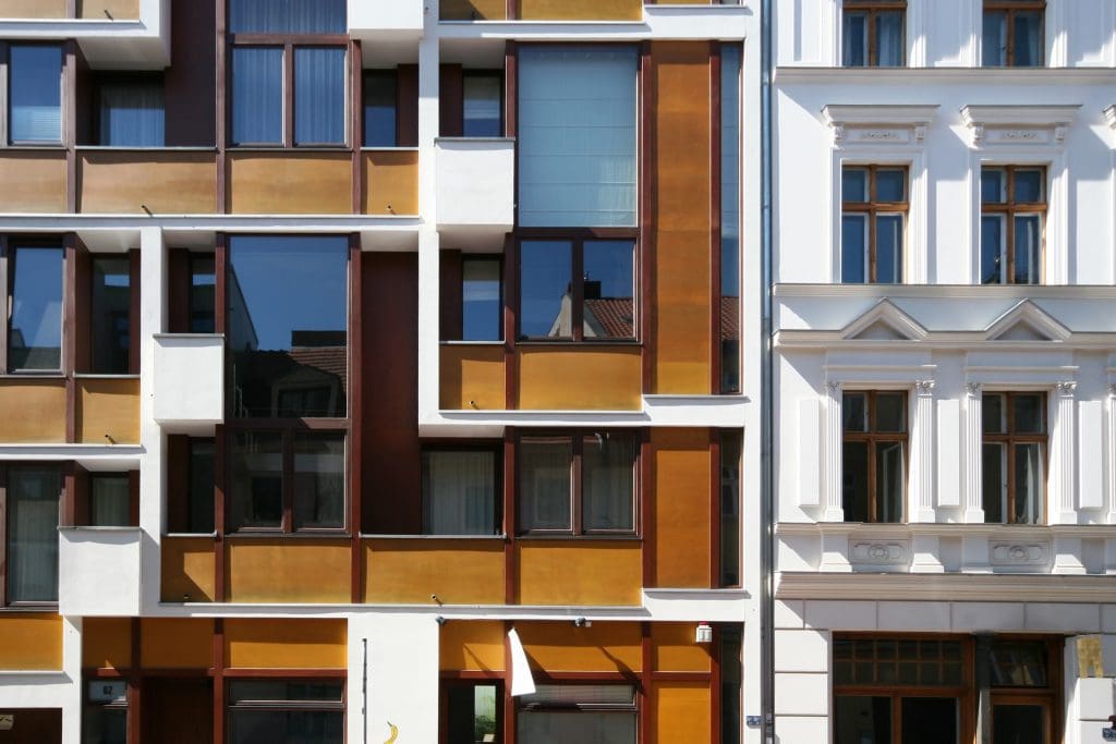 Két egymás mellett álló városi lakóépület homlokzata. A bal oldali épület modern stílusú, aszimmetrikus elrendezésű ablakaival, barna és aranyszínű burkolati elemekkel. A jobb oldali épület klasszikus, fehér homlokzattal, díszített párkányokkal és fa keretes ablakokkal.