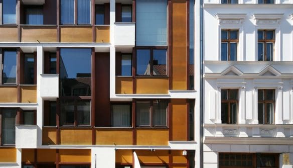 Két egymás mellett álló városi lakóépület homlokzata. A bal oldali épület modern stílusú, aszimmetrikus elrendezésű ablakaival, barna és aranyszínű burkolati elemekkel. A jobb oldali épület klasszikus, fehér homlokzattal, díszített párkányokkal és fa keretes ablakokkal.