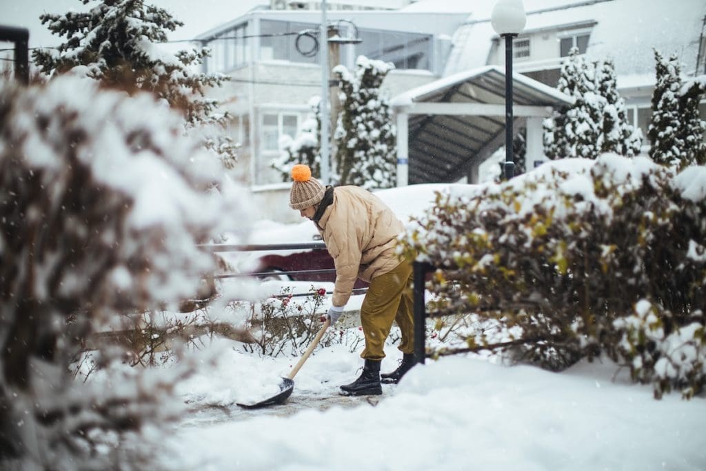 Téli ruhás személy havat lapátol egy udvaron, friss hóesésben, bokrok és házak között.