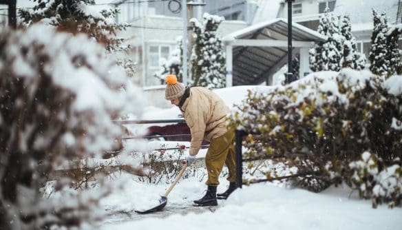 Téli ruhás személy havat lapátol egy udvaron, friss hóesésben, bokrok és házak között.