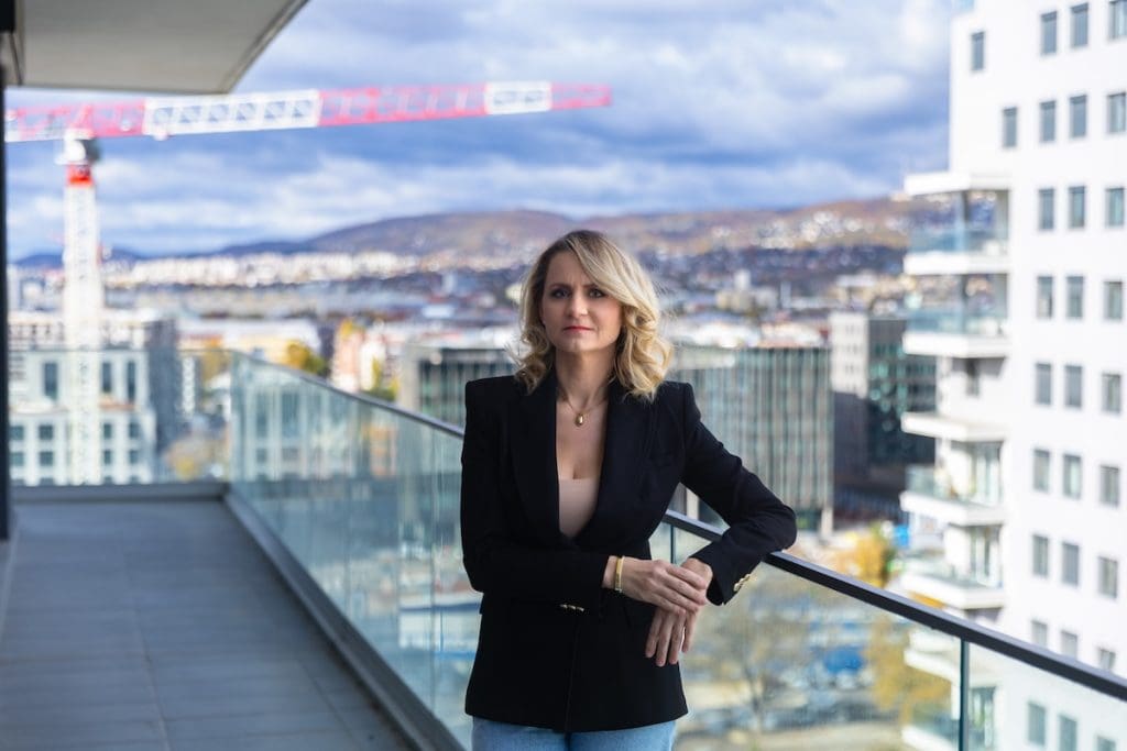 Zsellér Ilona ingatlanközvetítő portréja egy modern, új építésű ingatlan teraszán. Ilona elegáns fekete blézert visel, mögötte felhős ég és egy építkezés daruja látható.
