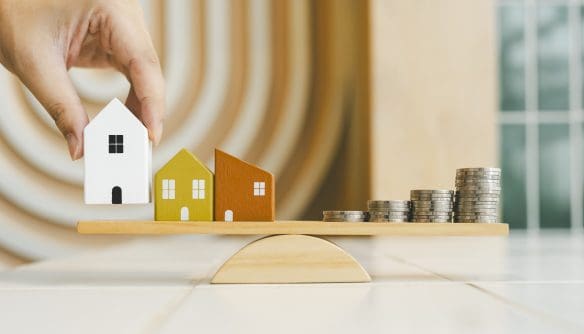 Egy letisztult, világos enteriőrben egy fa mérleg látható, amelynek bal oldalán három kis, stilizált faház áll egymás mellett. A házak különböző színűek: fehér, sárga és barna, egyszerű ablak- és ajtójelölésekkel. A bal szélső, fehér házat egy emberi kéz éppen felemeli vagy a mérlegre helyezi, finom, óvatos mozdulattal. A mérleg jobb oldalán egymásra rakott ezüstszínű érmék sorakoznak növekvő magasságban, ami az anyagi értéket vagy költségeket szimbolizálja. A mérleg enyhén kiegyensúlyozottnak tűnik, a házak és az érmék vizuálisan egymással „mérlegelésben” állnak. A háttér elmosódott, meleg tónusú, íves mintázatú fal- vagy dekorációs elem látható, amely nyugodt, modern hangulatot teremt. A kompozíció az ingatlanérték, lakhatás és pénzügyi döntések közötti egyensúlyt, illetve az ár–érték viszonyt szemlélteti.