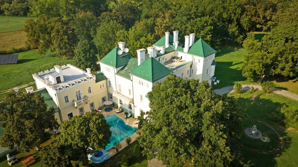 Légifelvételen egy nagyméretű, kastélyszerű épületegyüttes látható parkosított környezetben. Az épület világos, törtfehér–halványsárga homlokzattal rendelkezik, zöld színű, meredek tetőkkel és több, hangsúlyos toronyszerű kiemeléssel. A tetőn több fehér kémény és tetőfelépítmény látható, az épület architektúrája historizáló, reprezentatív jellegű. Az épület egyik belső udvarában szabálytalan formájú, világoskék színű úszómedence található napozóágyakkal és pihenőterületekkel. A kastélyt sűrű, zöld lombú fák veszik körül, nagy kiterjedésű, gondozott park és gyepfelületek ölelésében. A jobb oldalon kanyargó sétány és egy kör alakú díszkert vagy szökőkút is megfigyelhető. A környezet csendes, természetközeli, a háttérben erdős terület és nyílt zöld mezők látszanak. A felvétel napos időben készült, a hosszabb árnyékok kellemes, nyugodt hangulatot sugallnak, az ingatlan exkluzív, elszeparált jellegét hangsúlyozva.