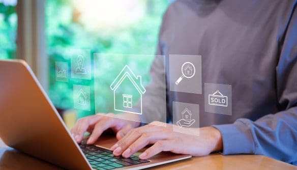Egy asztalnál ülő személy laptopon dolgozik, miközben a képernyő előtt lebegő, digitális ikonok jelennek meg az ingatlanpiac témájában. A grafikus elemek között látható egy ház szimbóluma, százalékjel, nagyítóval vizsgált ingatlan, „sold” (eladva) felirat és kézben tartott ház ikon. A vizuális elemek az online ingatlankeresést, -értékelést és -adásvételt jelképezik, modern, technológia-orientált környezetben. A kép az ingatlanpiac digitalizációját és az online döntéstámogatást hangsúlyozza.