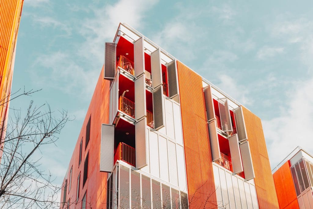 Modern, többemeletes társasház alulnézetből fotózva, narancssárga és világosszürke homlokzati elemekkel. Az épületen egymás fölött elhelyezkedő erkélyek láthatók, részben nyitható árnyékoló panelekkel és piros korlátokkal. A letisztult, kortárs építészeti stílusú lakóépület előtt egy lombtalan fa ága jelenik meg, a háttérben világoskék, enyhén felhős égbolttal.