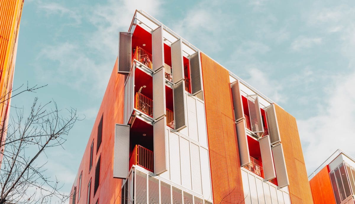 Modern, többemeletes társasház alulnézetből fotózva, narancssárga és világosszürke homlokzati elemekkel. Az épületen egymás fölött elhelyezkedő erkélyek láthatók, részben nyitható árnyékoló panelekkel és piros korlátokkal. A letisztult, kortárs építészeti stílusú lakóépület előtt egy lombtalan fa ága jelenik meg, a háttérben világoskék, enyhén felhős égbolttal.