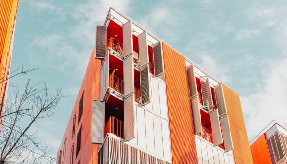 Modern, többemeletes társasház alulnézetből fotózva, narancssárga és világosszürke homlokzati elemekkel. Az épületen egymás fölött elhelyezkedő erkélyek láthatók, részben nyitható árnyékoló panelekkel és piros korlátokkal. A letisztult, kortárs építészeti stílusú lakóépület előtt egy lombtalan fa ága jelenik meg, a háttérben világoskék, enyhén felhős égbolttal.