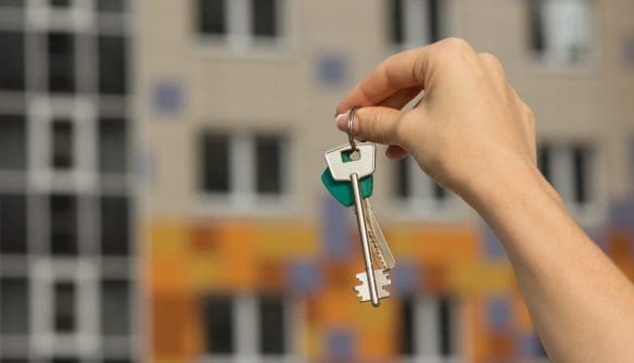 Egy női kéz lakáskulcsot tart a levegőben, a háttérben modern társasház homlokzata látható. A kép egy albérletárakkal, lakbérekkel kapcsolatos cikk illusztrációja.