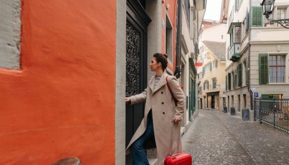 Albérlethez vagy rövid távú lakhatáshoz érkező nő egy régi városrész macskaköves utcájában: bőrönddel a kezében belép egy lakóépület kapuján. A kép a költözést, lakásbérlést, új otthonba érkezést és városi lakhatási helyzeteket illusztrálja.