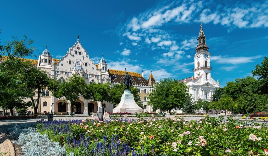 A kecskeméti városháza és katolikus templom, előtte zöld kerttel és virágokkal.