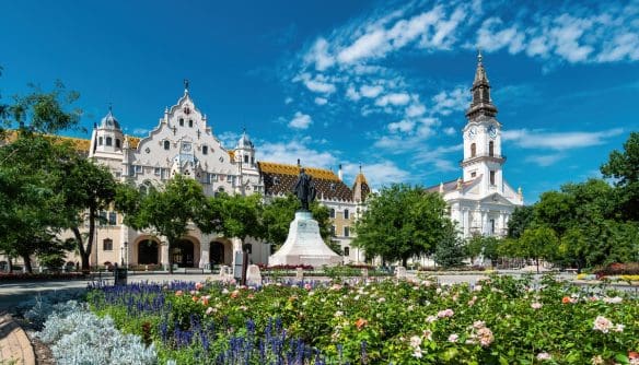 A kecskeméti városháza és katolikus templom, előtte zöld kerttel és virágokkal.