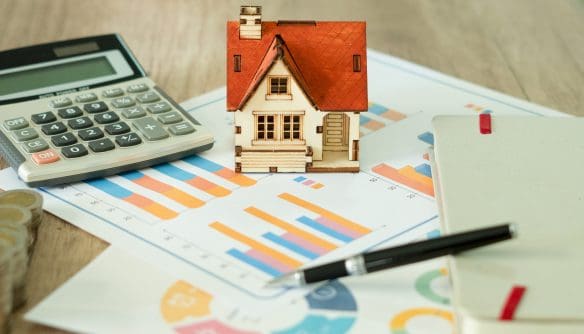 Lakásvásárlással és ingatlanpiaci elemzéssel kapcsolatos illusztráció: egy makett ház pénzügyi grafikonokat ábrázoló lapokon, mellette számológép, jegyzetfüzet és toll, az ingatlanárak és befektetések tervezésének szimbólumaként.
