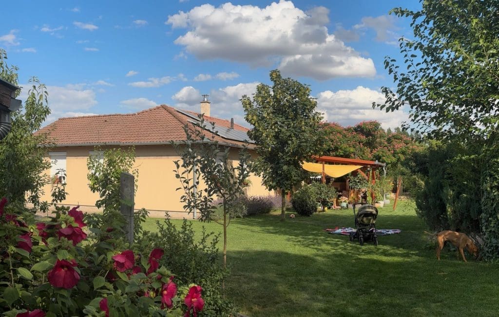 Családi ház tágas, zöld kerttel: gyep, fák és virágok között babakocsi és takaró, a háttérben terasszal és pergolával rendelkező, cseréptetős ház, nyugodt, kertvárosi környezetben.