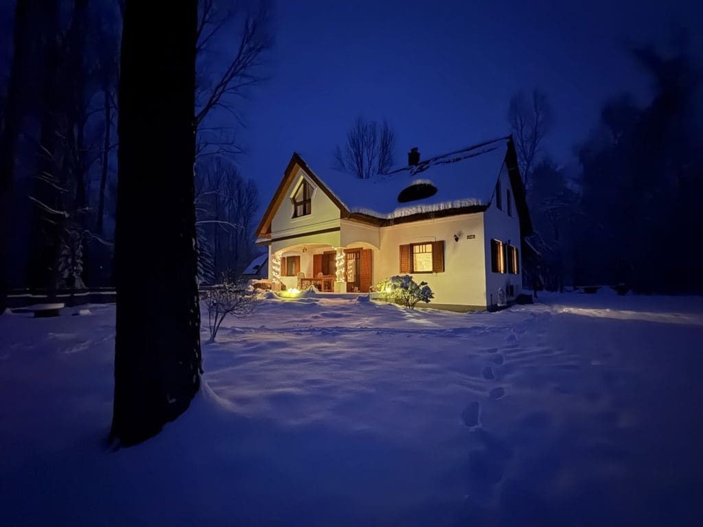Hóval borított kertben álló, meleg fényekkel kivilágított családi ház téli estében, a sötétkék ég alatt nyugodt, meghitt hangulattal.