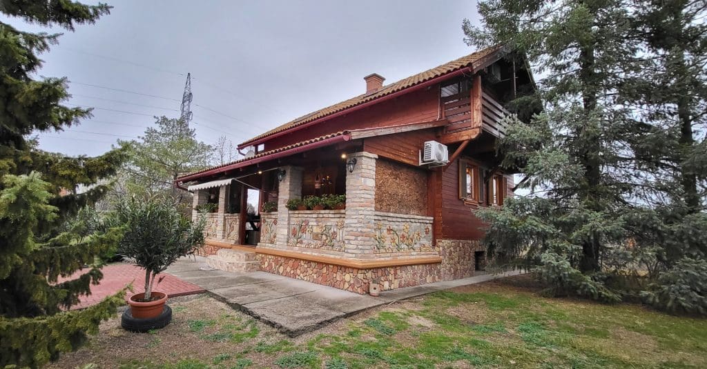 Fás kertben álló, rusztikus hangulatú fa–kő burkolatú családi ház fedett terasszal, emeleti erkéllyel és környező zöld növényzettel.