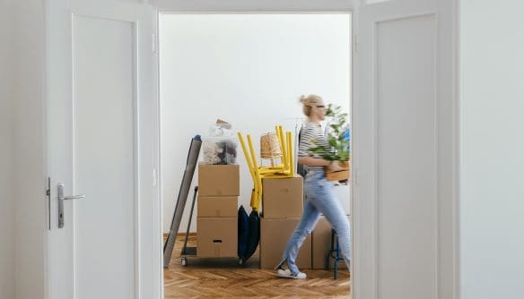 Nyitott ajtókereten keresztül egy világos szobába látni, ahol költözéshez előkészített kartondobozok és felpakolt bútorok állnak. Egy fiatal nő éppen átsétál a helyiségen, kezében növényt tart, miközben a parkettás padlón dobozok, felrakott sárga székek és egy molinóba tekert szőnyeg látható. A kép a költözés vagy új lakásba való beköltözés pillanatát ábrázolja.
