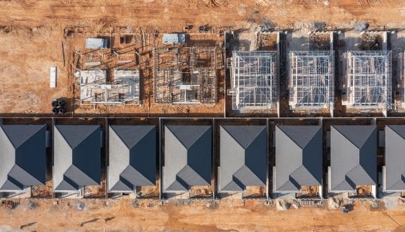 Felülnézeti, drónnal készült felvétel egy lakópark építéséről. Az alsó sorban már elkészült, egyforma tetős családi házak állnak egymás mellett rendezett telkeken, míg a felső részen több ingatlan még különböző építési fázisban van: láthatók alapozások, falazatok és tetőszerkezetek. A terület földes, építési anyagokkal és munkagépekkel, ami aktív kivitelezésre utal.