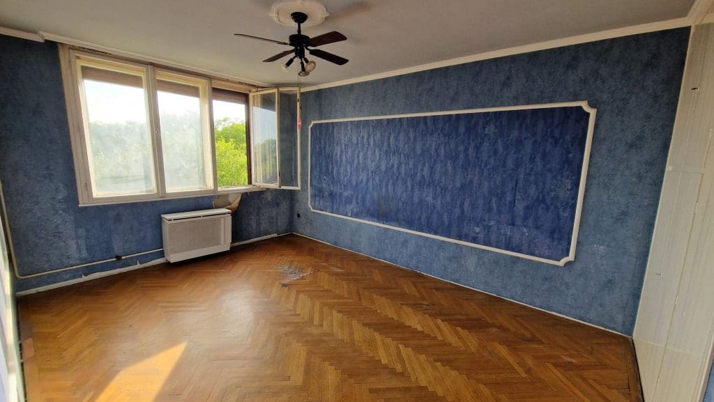 Üres, felújítandó szoba kék falakkal, halszálkamintás parkettával, mennyezeti ventilátorral és nagy ablakkal.
